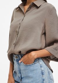 Osoba nosi voľnú taupe košeľu na gombíky zastrčenú do svetlomodrých vysokopásových džínsov s jednou rukou v vrecku.