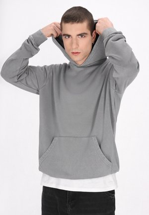 Młody mężczyzna poprawia kaptur swojej szarej bluzy, ma na sobie białą koszulę i czarne spodnie, na jednolitym białym tle.