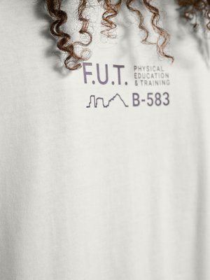 Close-up af hvid skjorte, der viser krøllet hår og trykt tekst "F.U.T. Physical Education & Training B-583" med lille bysilhuet grafik.