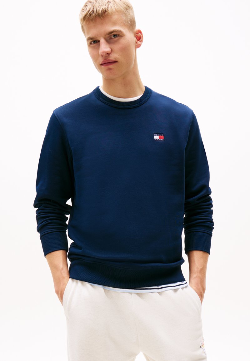 Navyblauwe sweatshirt met rib gebreide manchetten en halslijn, voorzien van een kleine logo patch met rode en witte accenten op de borst. Zachte stoftextuur.