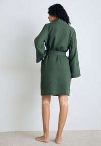 Robe vert à texture en nid d'abeille avec des manches larges, ceinturée à la taille, arrivant au genou, vue de dos. Pieds nus sur un tapis clair.