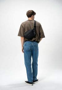Borsa a tracolla nera con chiusura a zip e tracolla regolabile, indossata sopra una maglietta oversized marrone e jeans di denim blu.