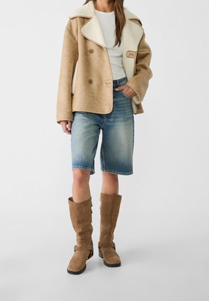 Femme portant une veste en peau retournée beige, un haut blanc, un short en jean longueur genou et des bottes hautes en daim beige, debout devant un fond uni.