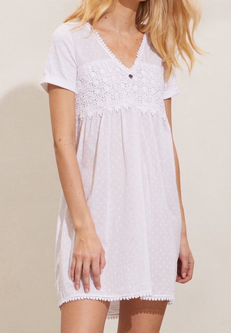 Femme portant une robe blanche courte avec des pois texturés, un panneau en dentelle florale et des manches courtes, se tenant devant un arrière-plan uni.