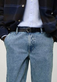 Hombre vestido con una camisa de botón azul claro, chaqueta de cuadros oscuros, jeans azul claro y cinturón negro, con las manos en los bolsillos.