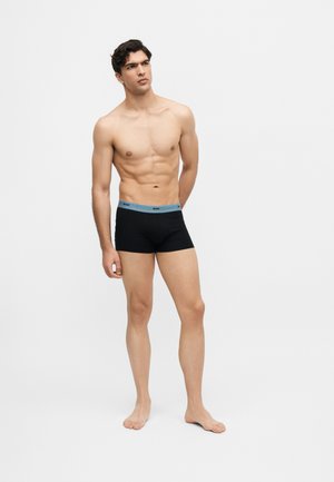 Homme musclé debout pieds nus portant un boxer noir avec une ceinture bleue de la marque "BOSS", regardant légèrement vers sa gauche.