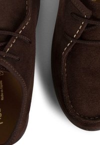 Hnědé semišové boty s kulatým tvarem, s viditelným prošíváním a šněrováním. Měkká textura a minimalistický styl.