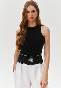 Czarny prążkowany top na ramiączkach o dopasowanym fasonie. Posiada kontrastowy, biały pas z logo. Gładka struktura, zestawiona z jasnobeżowymi spodniami.