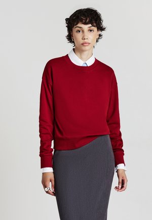 Felpa rossa con scollatura a girocollo, maniche lunghe e polsini a costine, abbinata a una gonna grigia texture. Una camicia bianca con colletto è visibile sotto.