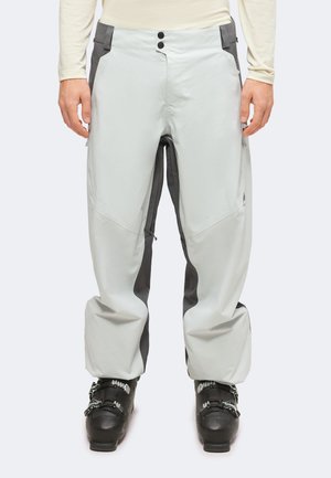 Persona con pantalones de esquí gris claro y gris oscuro y botas de esquí negras, de pie recto con los brazos relajados a los lados.