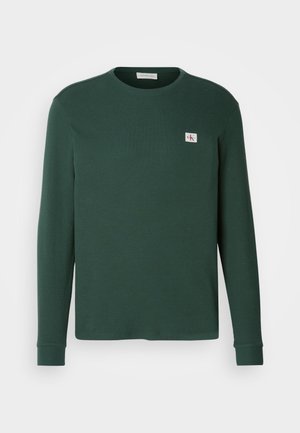 Pulover verde cu mânecă lungă, realizat dintr-o țesătură cu dungi, cu guler rotund. Prezintă o mică eticheta cu logo pe piept și manșete cu dungi.