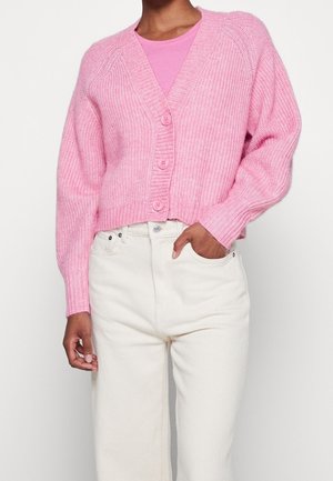 Osoba w różowym rozpinanym swetrze na guziki, nosząca różowy top, w połączeniu z jasnobeżowymi, wysokim stanem, nogawkami, z ręką w kieszeni, na białym tle.