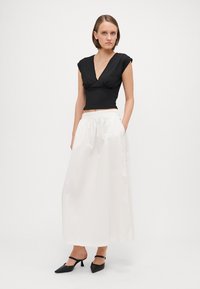 Haut noir ajusté à manches courtes, associé à une longue jupe blanche fluide ; le modèle porte des chaussures noires à talons pointus avec des brides autour des chevilles.