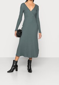 Kvinna iklädd en långärmad, V-ringad, ribbad grön midiklänning, matchad med svarta klackiga ankelstövlar, hållande en svart kuvertväska.