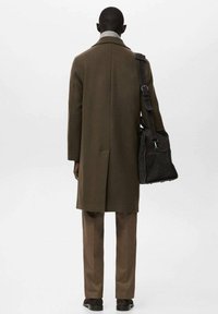 Abrigo de lana verde oliva con una sola abertura trasera, combinado con pantalones marrones. Un bolso mensajero negro cuelga de un hombro.