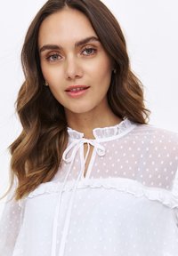 Biała, przezroczysta bluzka z wzorem w kropki, z falbaniastym dekoltem, wiązanym zapięciem i miękką teksturą. Podkreślona delikatnymi falbanami przy szwie klatki piersiowej.