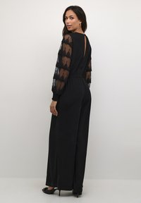 Schwarzer Jumpsuit mit durchsichtigen, gestuften schwarzen Spitzeärmel und V-Ausschnitt auf der Rückseite. Weit geschnittene Silhouette, glattes Material und minimaler Hardware.
