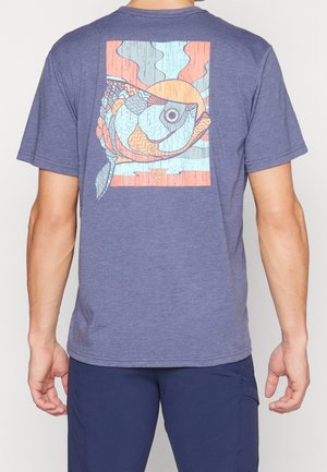 Personne portant un t-shirt bleu avec un design abstrait coloré de poisson et un motif de rayons de soleil dans le dos, associé à un short bleu marine.