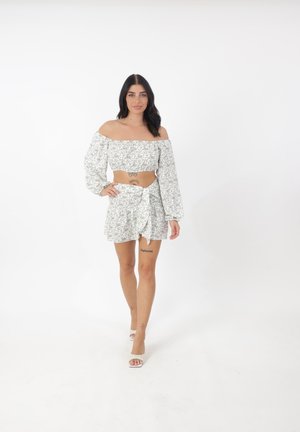 Weißes Off-Shoulder-Crop-Top mit Puffärmeln, kombiniert mit einem gemusterten Rock mit vorderem Bindeband. Blumenmuster in Grün und Beige.