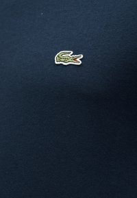 Σκούρο μπλε ύφασμα με μικρό κεντημένο πράσινο λογότυπο κροκοδείλου με ανοιχτό κόκκινο στόμα, τοποθετημένο στο κέντρο του υλικού.