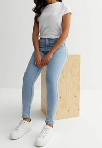 Jeans ajustados de color azul claro combinados con una camiseta blanca, acompañados de zapatillas blancas. Modelo sentado sobre un pedestal de madera clara.