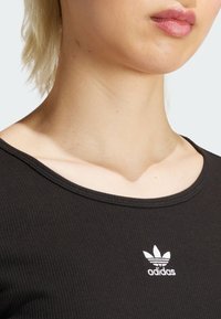 Juoda linkiuota marškinėliai su plačiu iškirpimu, su mažuoju baltu siuvinėjimu Adidas logotipu kairėje pusėje. Lygus paviršius.