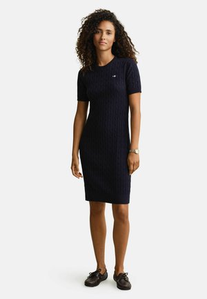 Vrouw draagt een marineblauwe kabelgebreide jurk met korte mouwen en bruine leren veterlaarzen, met een horloge om haar linkerpols.