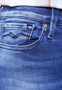 Jeans de mezclilla azul con un primer plano de un bolsillo trasero que presenta un distintivo patrón de costura en zigzag y un detalle de botón de metal.