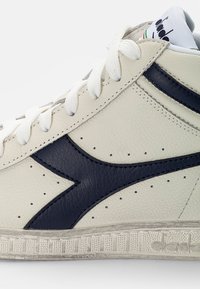 Baskets montantes blanches en cuir avec des accents navy, surface texturée et design perforé. Elles disposent de lacets blancs et d'une semelle en caoutchouc à motifs.