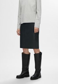 Zwarte leren kniehoge laarzen met een vierkante neus en een dikke rubberen zool, gecombineerd met een grijs langalarm shirt en een donkere rok.