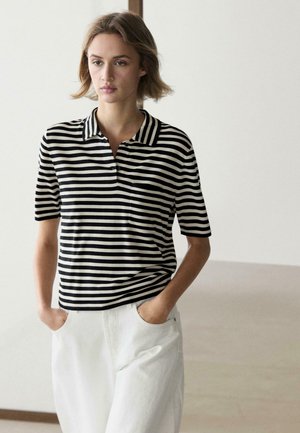 Donna con capelli corti che indossa una polo a maniche corte a righe bianche e nere e pantaloni bianchi con le mani nelle tasche.