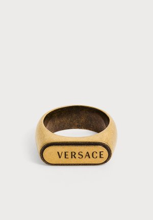 Arany tónusú fém gyűrű, négyszögletes felülettel, amelybe az "VERSACE" szó nagybetűkkel van gravírozva.