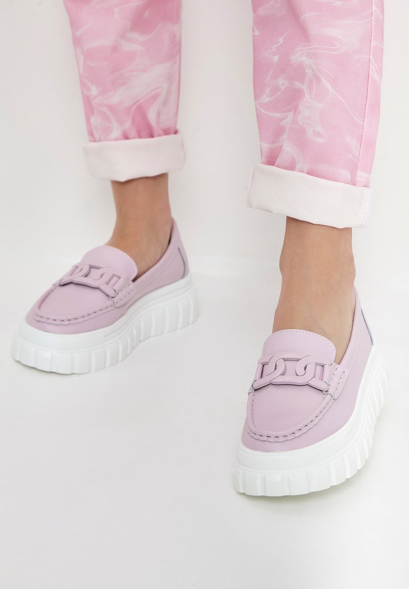 Mocasini violet deschis cu talpă albă groasă, având o accentuare decorativă cu lanț și detalii cusute. Purtați cu pantaloni roz cu model marmorat, răsuciți la glezne.