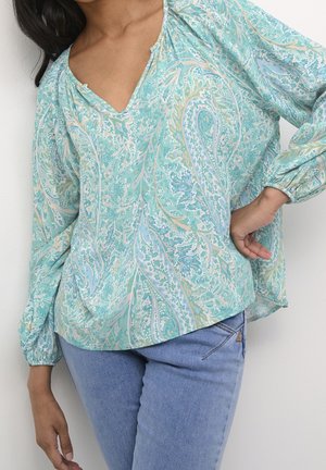 Vrouw die een lichtblauw paisley blouse met lange mouwen en elastische manchetten draagt, gecombineerd met lichtblauwe jeans, poseert met één hand op haar heup.