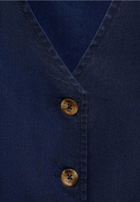 Navy-blaues Denim-Hemd mit V-Ausschnitt, drei braunen Knöpfen und sichtbaren Nähten entlang der Knopfleiste und Kragenkanten.