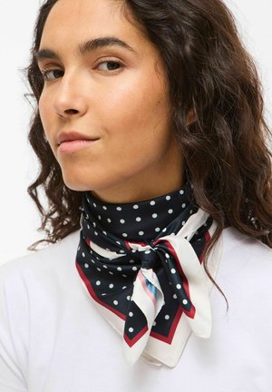 Jauna sieviete ar cirtainiem tumšiem matiem, valkājot melnu lakatiņu ar baltām un sarkanām malām un baltiem punktiem, kopā ar baltu krekliņu.