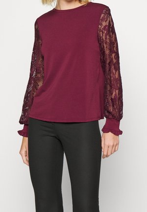 Haut bordeaux avec un corps en tissu doux et des manches longues en dentelle. Les manches présentent un motif floral et des poignets froncés. Assorti avec un pantalon noir.