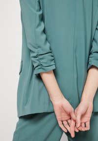 Teal blouse met geplooide mouwboorden en zijnaden. De stof lijkt glad en lichtgewicht, wat zorgt voor een ontspannen pasvorm. Handen samengevouwen achter de rug.