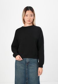 Jeune femme avec des cheveux raides, portant un sweat à manches longues noir et une jupe en denim bleue, se tenant devant un fond blanc.