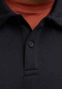 Sort poloshirt med tekstureret vævning, der har en enkelt knapplade og en synlig rød undertrøjes halsudskæring nedenunder.