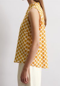 Mujer lleva una blusa sin mangas de color mostaza y blanco con un patrón geométrico, de corte holgado y con cuello alto, combinada con pantalones de color claro.