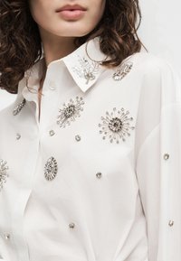 Γυναίκα που φοράει λευκό πουκάμισο με κουμπιά διακοσμημένο με ασημένια κοσμήματα σε σχήματα εκρήξεων αστεριών και κλαδιών.