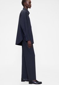 Donkergrijs oversized shirt en wijde broek van een getextureerde stof, gecombineerd met zwarte schoenen. Voorzien van knoopmanchetten en zijsplitten.