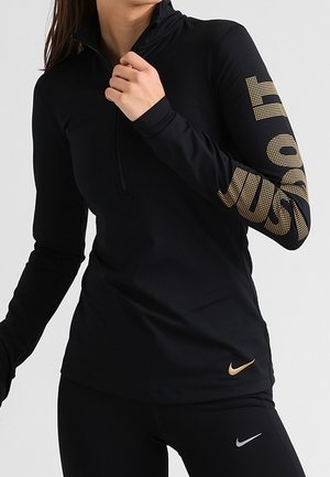 Frau trägt ein schwarzes langärmliges Sportoberteil mit goldfarbenem "JUST DO IT"-Text am Ärmel und einem goldfarbenen Nike-Logo in der Nähe des Saums.