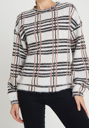 Femme portant un pull blanc avec un motif à carreaux noir et marron et un pantalon noir, debout avec une main détendue à ses côtés.