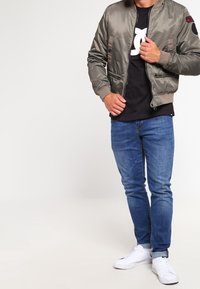 Olivově zelená bomber bunda se zipem vpředu, bočními kapsami a žebrovanými manžetami, v kombinaci s černým tričkem a modrými džíny. Bílé tenisky.