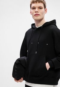 Sudadera negra con bolsillo canguro, detalles en plata en los cordones y un logo minimalista. El modelo sostiene un objeto negro mientras la lleva puesta.