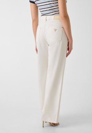 Back view of person wearing high-waisted white wide-leg jeans, striped shirt, and gold high-heeled sandals against a plain background.