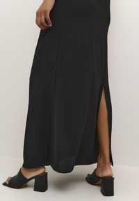 Black maxi dress with side slit, made of lightweight fabric. Paired with black block-heeled mules, showing smooth texture and sleek design.