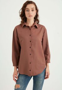 Camicia marrone con bottoni, colletto, maniche lunghe e orlo arrotondato. Il materiale sembra liscio; presenta bottoni scuri lungo il davanti.
